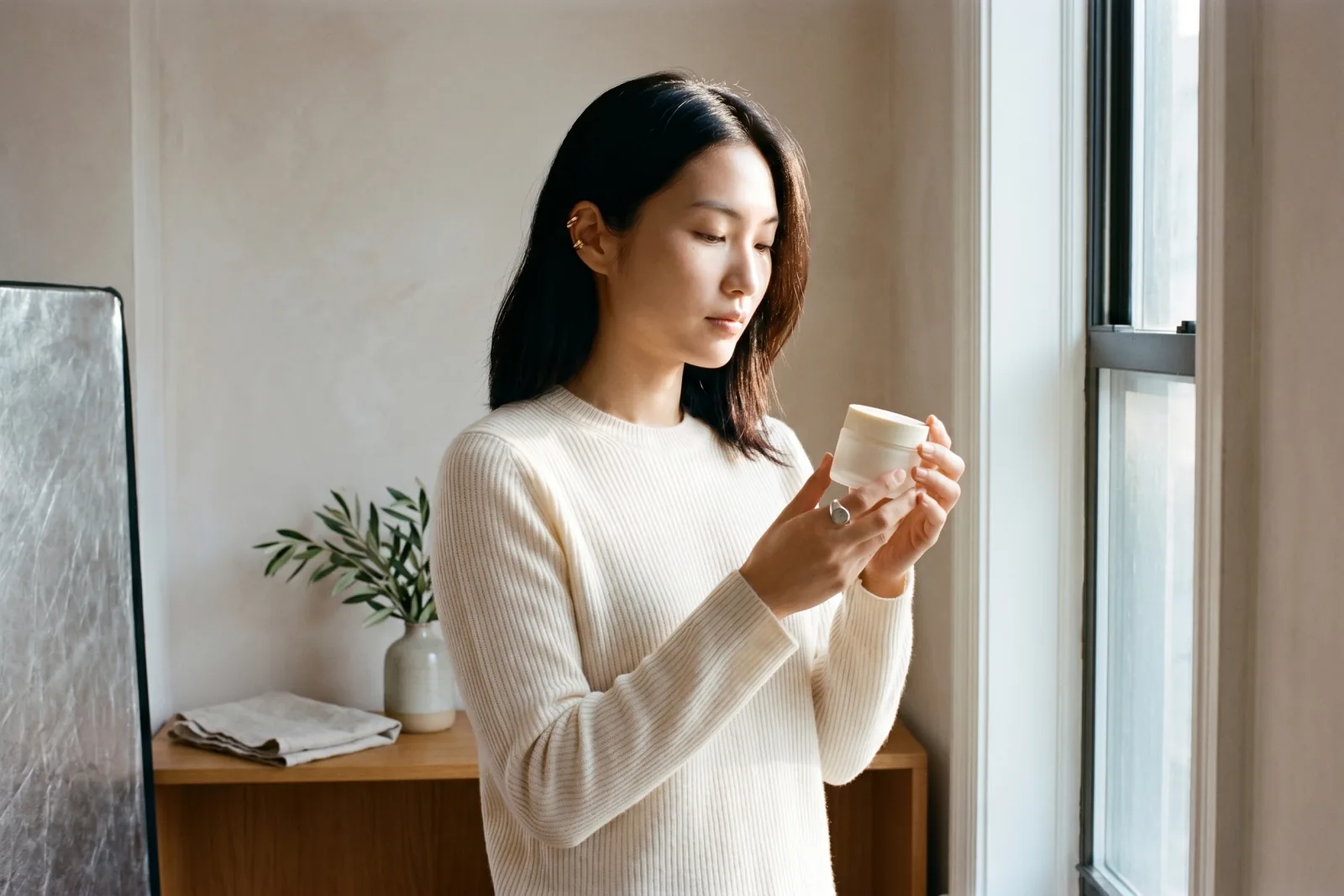 Editorial brand-shoot photograph of a Playcut custom AI model at a sunlit studio window holding a frosted-glass skincare jar, brushed-steel reflector at edge of frame, freeze-frame of a campaign video