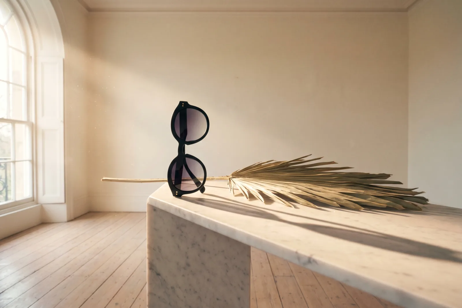 Theatrical editorial product photograph of unbranded oval-frame matte black acetate sunglasses on a Carrara marble plinth in an empty cream gallery room, dried palm leaf casting a long dramatic afternoon shadow