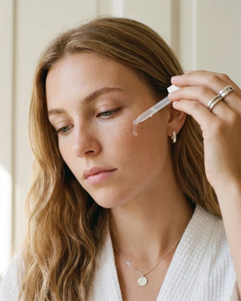 Editorial beauty portrait of Sienna, a young honey-blonde lifestyle ambassador with freckles applying a serum drop from a frosted-glass dropper, white waffle bathrobe, soft natural daylight, Playcut Actor Engine generation